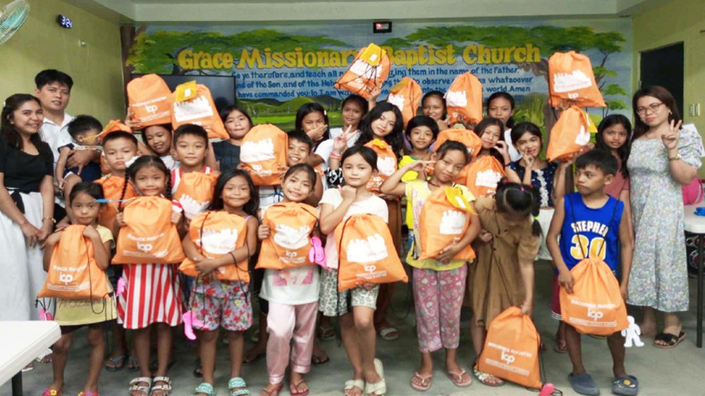Children at a church holding orange ICP backpacks filled with hygiene and school supplies.